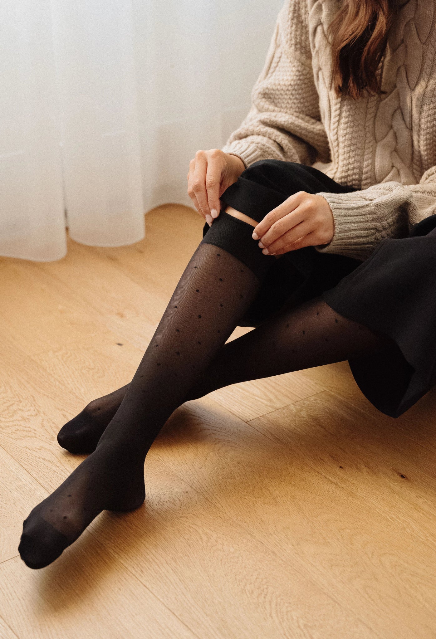 Femme assise sur parquet enfilant les mi-bas Polka Glow compression légère noir – Motif pois – Soje Lingerie